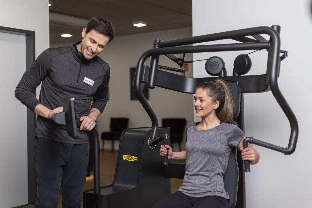 Sporttherapeut begleitet eine Frau beim Gerätetraining und überwacht die Einstellungen