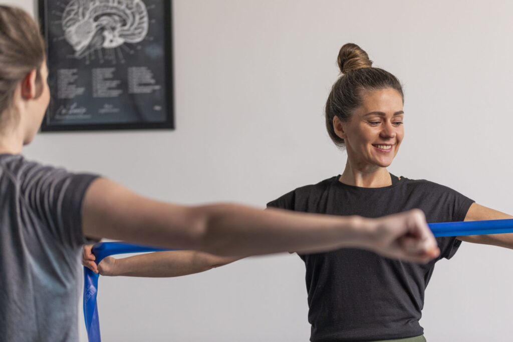 Zwei Frauen führen gemeinsam eine Übung mit einem blauen Theraband durch, eine von ihnen lächelt dabei