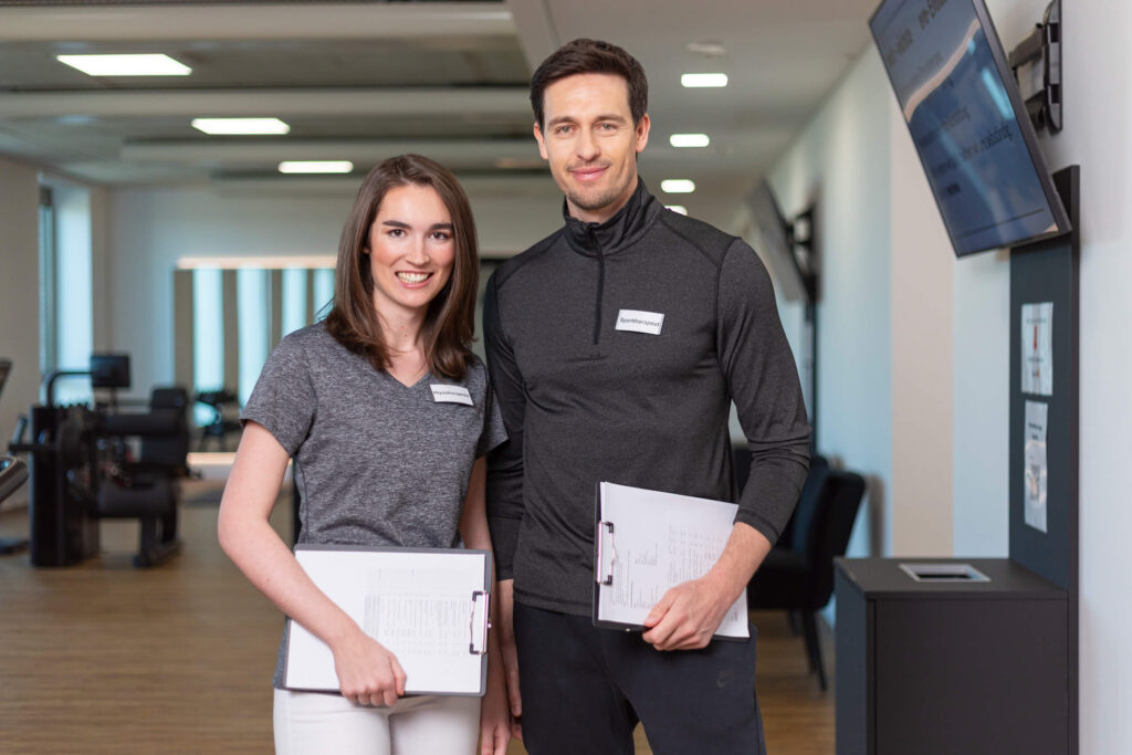Zwei Mitarbeitende in sportlicher Kleidung mit Klemmbrettern in einem Trainingsbereich